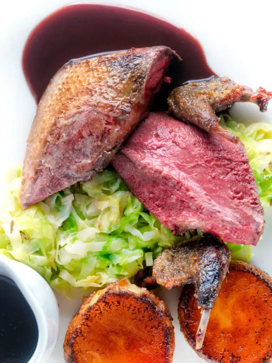 Overhead close-up roast pigeon breasts, confit legs, red wine sauce, buttered cabbage and fondant potatoes.