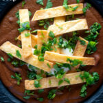 Overhead spicy vegan black bean soup topped with crispy tortillas and fresh coriander featuring a title overlay.