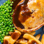 Overhead close-up steak and Stilton pie with chips, peas and gravy.