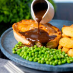 Rich gravy being poured over a steak and stilton pie served with chips and garden peas.