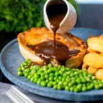 Rich gravy being poured over a steak and stilton pie served with chips and garden peas.
