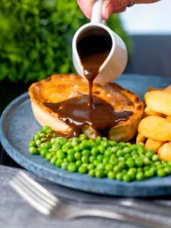 Rich gravy being poured over a steak and stilton pie served with chips and garden peas.