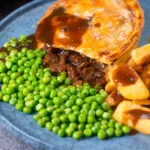 Cut open steak and stilton pie showing filling served with chips and garden peas.