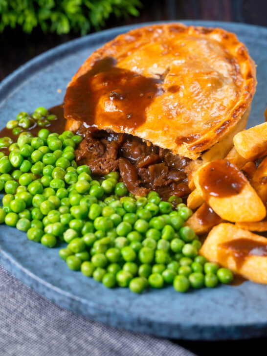 Cut open steak and stilton pie showing filling served with chips and garden peas.