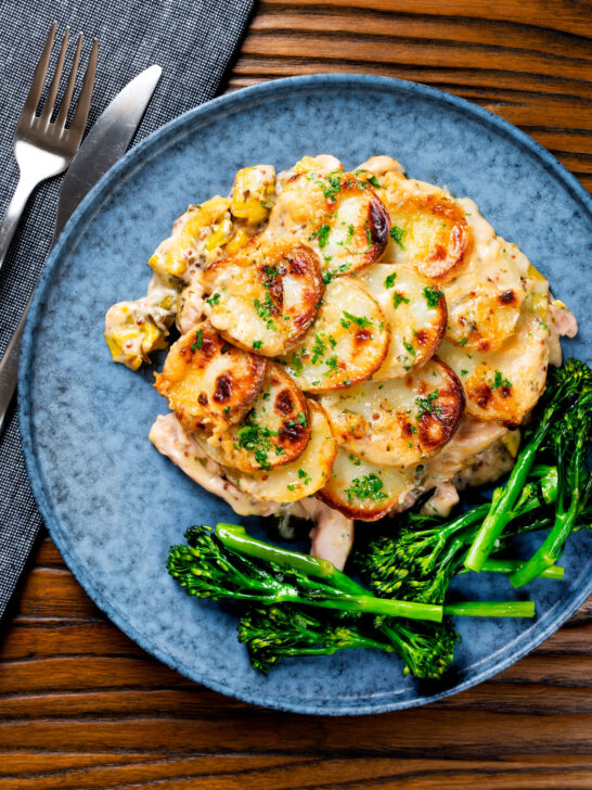 Overhead scalloped potato topped creamy chicken and leek hotpot with roasted tenderstem broccoli.