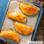 Overhead homemade cheese and onion pasties in shortcrust pastry on a baking sheet featuring a title overlay.