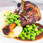 Gravy being poured over minted lamb shanks served with peas and mashed potato.