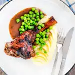 Overhead oven cooked minted lamb shanks served with peas, mashed potato and red wine gravy featuring a title overlay.