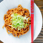 Overhead vegan tofu noodle stir fry with peanuts and dan dan sauce garnished with spring onions.