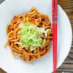 Overhead vegan tofu noodle stir fry with peanuts and dan dan sauce garnished with spring onions featuring a title overlay.