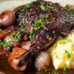 Close up coq au vin, chicken legs braised in red wine with vegetables and bacon featuring a title overlay.