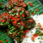 Overhead close-up beef rendang curry with rice, chilli and coriander served on a banana leaf.