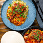 Overhead duck leg tagine with Moroccan flavours served with buttered couscous and fresh coriander featuring a title overlay.