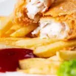 Close-up chip shop fried fish with crispy batter opened to show moist haddock featuring a title overlay.