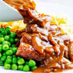 Onion gravy being poured over a piece of Cumberland sausage ring with peas and mash.