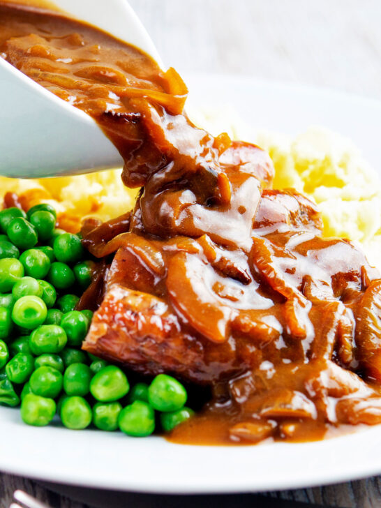 Onion gravy being poured over a piece of Cumberland sausage ring with peas and mash.