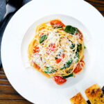 Overhead roasted garlic spaghetti with tomatoes and spinach garnished with parmesan cheese.