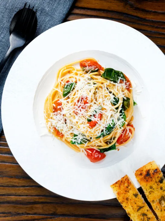 Overhead roasted garlic spaghetti with tomatoes and spinach garnished with parmesan cheese.