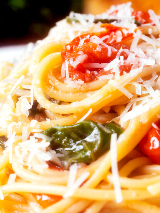 Close-up roasted garlic spaghetti with tomatoes and spinach garnished with parmesan cheese.