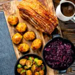 Overhead slow roast pork belly with crackling, roast potatoes, sprouts, red cabbage and gravy featuring a title overlay.