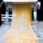 Homemade duck egg pasta being cut into tagliatelle in a pasta machine featuring a title overlay.