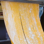 Close-up homemade duck egg pasta being cut into tagliatelle in a pasta machine.