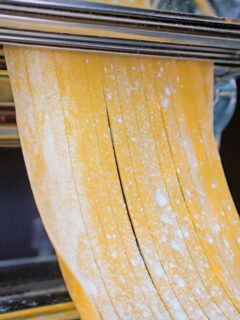 Close-up homemade duck egg pasta being cut into tagliatelle in a pasta machine.