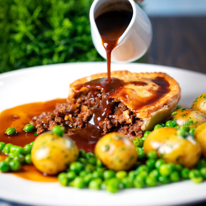 Individual British minced beef and onion pie with shortcrust pastry served with peas and potatoes.