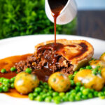 Gravy being poured over a homemade minced beef and onion pie served with peas and potatoes.