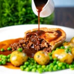 Gravy being poured over a homemade minced beef and onion pie served with peas and potatoes.