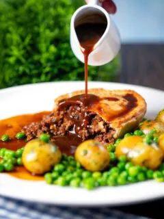 Gravy being poured over a homemade minced beef and onion pie served with peas and potatoes.