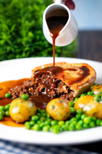 Gravy being poured over a homemade minced beef and onion pie served with peas and potatoes.