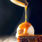 Hot caramel sauce being poured over British sticky toffee pudding and vanilla ice cream.