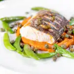 Baked or roasted hake with romesco sauce, capers and sugar snap peas featuring a title overlay.