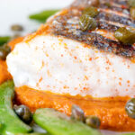 Close-up baked or roasted hake with romesco sauce, capers and sugar snap peas featuring a title overlay.