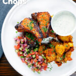 Overhead tandoori lamb chops with roasted Bombay potatoes, raita and kachumber salad, featuring a title overlay.