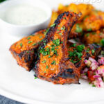 Tandoori lamb chops with roasted Bombay potatoes, raita and kachumber salad, featuring a title overlay.