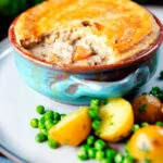 Rabbit pot pie with a suet crust cut open to show creamy filling served with potatoes and peas.