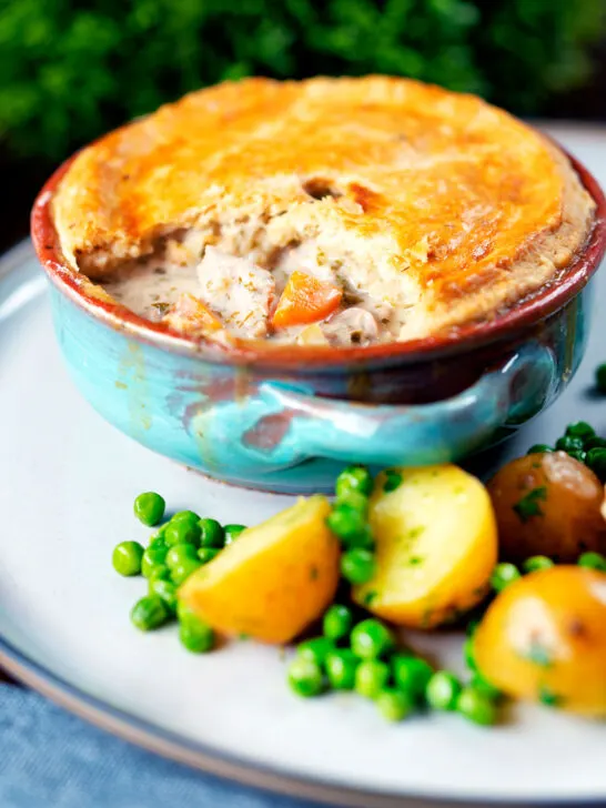 Rabbit pot pie with a suet crust cut open to show creamy filling served with potatoes and peas.