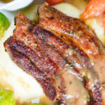 Overhead close-up crispy roast pork belly slices with a cider sauce, mashed potato and veggies.