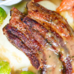 Overhead close-up crispy roast pork belly slices with a cider sauce, mashed potato and veggies.