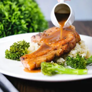 Sauce being poured over brown sugar pork chops served with celeriac mash and broccoli.