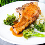 Sauce being poured over brown sugar pork chops served with celeriac mash and broccoli.