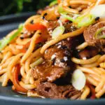 Close-up beef yakisoba noodle stir fry garnished with sesame seeds and spring onions.