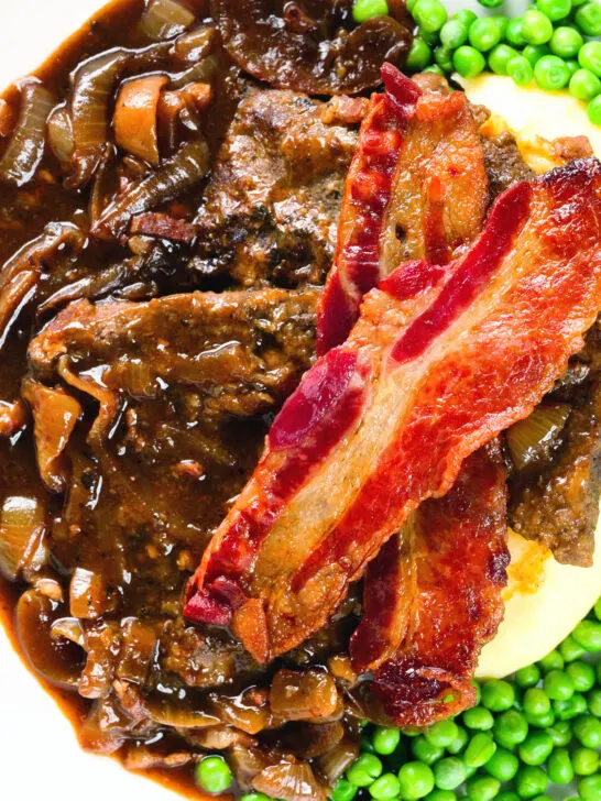 Overhead close-up liver and onions, in onion gravy served with crispy bacon, mash and peas.