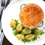 Overhead pork and apple pie with a puff pastry lid served with buttered new potatoes and peas.
