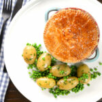 Overhead pork and apple pie with a puff pastry lid served with buttered new potatoes and peas.