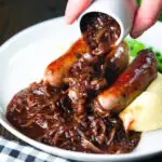 Thick onion gravy being poured over bangers (sausages) and mash.