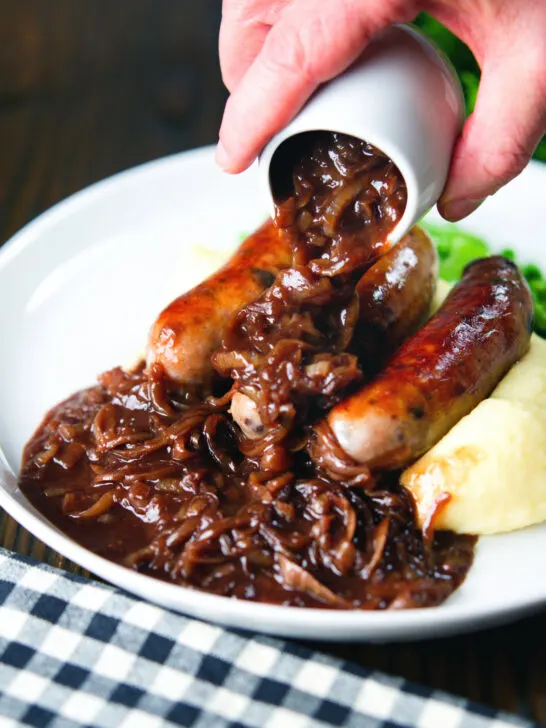 Thick onion gravy being poured over bangers (sausages) and mash.