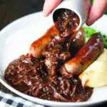 Thick onion gravy being poured over bangers (sausages) and mash .